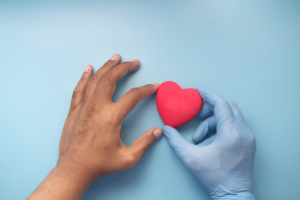 Two hands holding a red heart: one hand has brown skin and the other is in a blue medical glove